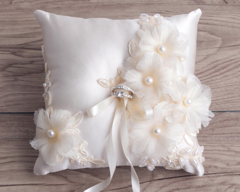 May include: A white satin ring pillow with delicate lace and pearl embellishments. Two silver rings are placed on the pillow.