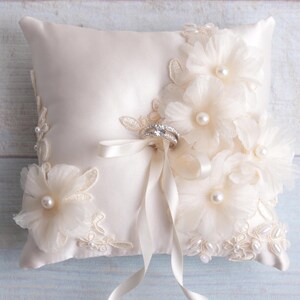 May include: A white satin ring pillow with delicate lace trim and pearl embellishments. The pillow is adorned with three large, white fabric flowers and a satin ribbon.