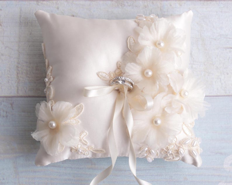 May include: A white satin ring pillow with delicate lace trim and pearl embellishments. The pillow is adorned with three large, white fabric flowers and a satin ribbon.