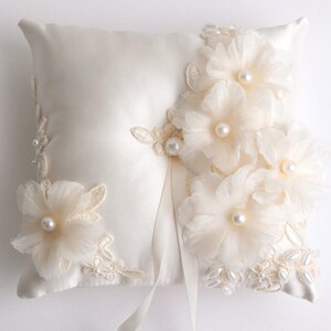 May include: A white satin ring pillow with delicate lace and pearl embellishments. The pillow is adorned with ivory fabric flowers and pearls.