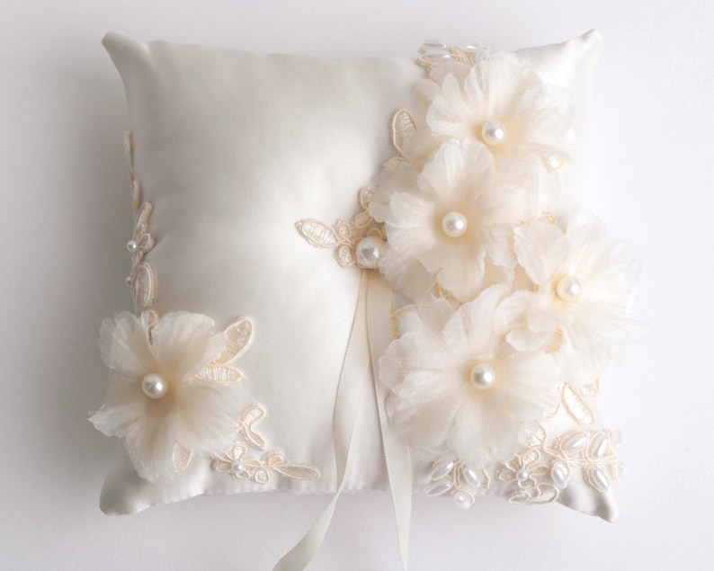 May include: A white satin ring pillow with delicate lace and pearl embellishments. The pillow is adorned with ivory fabric flowers and pearls.