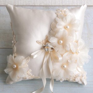 May include: A white satin ring pillow with delicate lace trim and pearl accents. The pillow is adorned with three large, white fabric flowers. Two rings are placed on the pillow, secured with a white satin ribbon.