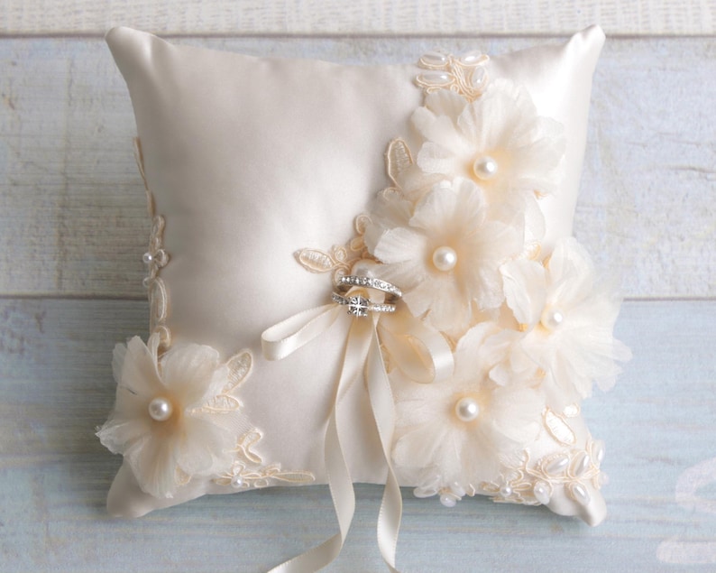 May include: A white satin ring pillow with delicate lace trim and pearl accents. The pillow is adorned with three large, white fabric flowers. Two rings are placed on the pillow, secured with a white satin ribbon.