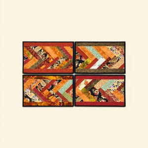 May include: Four rectangular quilted placemats with a black border. Each placemat features a chevron pattern in shades of orange, red, yellow, and green, with floral and paisley fabric accents. The placemats are arranged on a cream-colored surface.