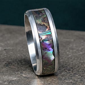 May include: A silver ring with a rectangular abalone inlay. The inlay features iridescent colors of green, purple, and blue. The ring has a polished finish and a modern design.