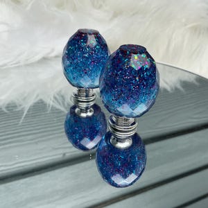 May include: Two decorative cabinet knobs with a faceted, oval design. The knobs feature a gradient of blue and purple glitter encased in clear resin, mounted on a silver-toned base. The knobs are displayed on a reflective surface.