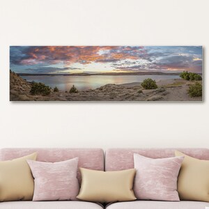 May include: A panoramic photograph of a lake with a cloudy sky and a pink and orange sunset. The lake is surrounded by trees and rocks.