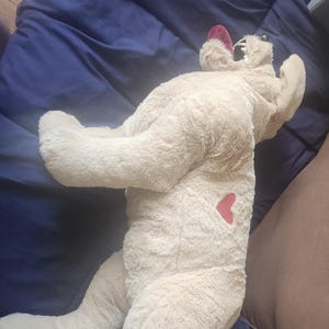 May include: A plush, beige teddy bear with a red heart on its side and red accents on its ears. The stuffed animal is lying on a blue blanket, with a brown pillow and a white object in the background.