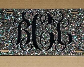 May include: Glitter license plate with a black monogram. The plate has a sparkly, multi-colored glitter finish. The monogram features the letters BCL in a decorative, intertwined font. The plate has four mounting holes.
