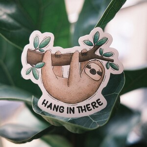 May include: A white sticker with a cartoon illustration of a brown sloth hanging from a branch with green leaves. The text "HANG IN THERE" is printed below the sloth.