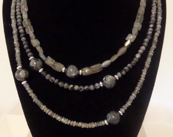May include: A layered necklace with three strands of dark gray and light gray beads. The necklace is made of various sizes and shapes of beads, including round, oval, and rectangular beads. The necklace is displayed on a black velvet mannequin.