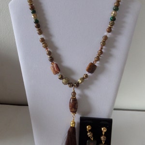 May include: A brown and gold beaded necklace with a tassel pendant and matching earrings. The necklace features a variety of brown, gold, and green beads, including a large brown bead with a gold accent. The earrings are made of brown and gold beads and have a square shape.