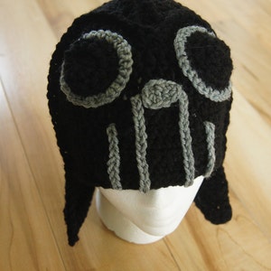 May include: Black crocheted hat with gray accents, resembling a face with two large eyes. The hat has long ear flaps.