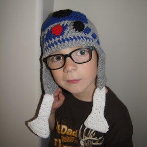May include: A child wearing a grey and blue crocheted hat with white ear flaps. The hat features a red and black design resembling a robot. The child is also wearing black framed glasses and a brown t-shirt with a deer graphic and the text "My Dad Can Hunt You're Not Bad!"