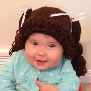 May include: A baby wearing a brown crocheted hat with white ribbons and a blue and white polka dot shirt.