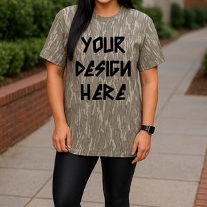 Puede incluir: Camiseta de manga corta con estampado de camuflaje en tonos verdes y marrones. El texto "YOUR DESIGN HERE" está en negro. Las palabras "MOSSY OAK BOTTOM LAND" están impresas en la parte superior de la imagen. La persona lleva leggings negros.