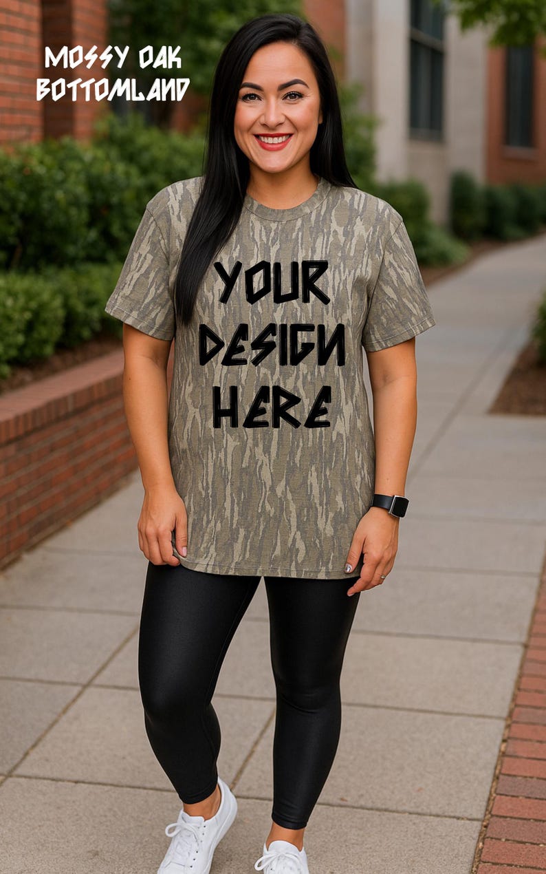 Puede incluir: Camiseta de manga corta con estampado de camuflaje en tonos verdes y marrones. El texto "YOUR DESIGN HERE" est&aacute; en negro. Las palabras "MOSSY OAK BOTTOM LAND" est&aacute;n impresas en la parte superior de la imagen. La persona lleva leggings negros.