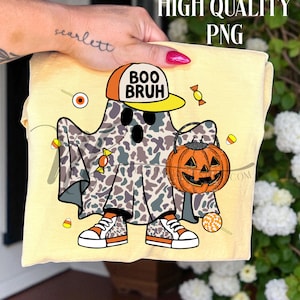 May include: A light yellow t-shirt featuring a cartoon ghost in a camouflage sheet, wearing a hat that says "BOO BRUH". The ghost is holding a jack-o'-lantern bucket and wearing sneakers. Candy corn and other candies are scattered around the ghost.