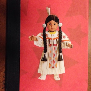 May include: A red notebook with a black spine and a white doll wearing a white dress with colorful embroidery and a feather headdress. The doll has long black braids and brown eyes.