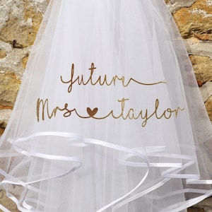 Pode incluir: Um véu de noiva branco com várias camadas e uma borda de fita. O véu tem o texto "Future Mrs. Taylor" em letras douradas. O véu está pendurado em uma parede de pedra.