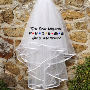 Könnte beinhalten: Ein weißer Brautschleier mit mehreren Lagen und einer Borte aus Bändern. Der Text "THE ONE WHERE PHOEBE GETS MARRIED" ist in Schwarz gedruckt, wobei das Wort "PHOEBE" im Stil der Fernsehserie "Friends" gehalten ist. Der Schleier hängt an einer Steinmauer.