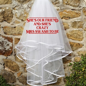 Może przedstawiać: Biały welon ślubny z wieloma warstwami i białym wykończeniem z wstążki. Welon ma czerwony napis "SHE'S OUR FRIEND AND SHE'S CRAZY MRS SWASH TO BE". Welon wisi na kamiennej ścianie.