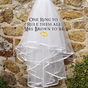 Może przedstawiać: Biały welon ślubny z warstwowym tiulem i wykończeniem z satynowej wstążki. Welon ma napis "One Ring to Rule Them All Mrs Brown to Be" i złotą grafikę pierścienia.