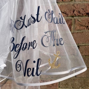 May include: White bridal veil with the text "The Last Sail Before The Veil" in navy blue script. A gold glitter anchor is also featured on the veil. The veil has a white trim.