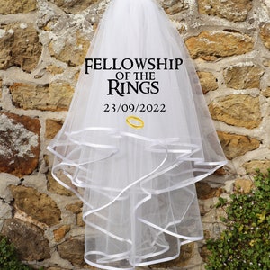 Może przedstawiać: Biały welon ślubny z wieloma warstwami i białym wykończeniem z wstążki. Na welonie wydrukowano napis "FELLOWSHIP OF THE RINGS", a także datę "23/09/2022" i grafikę złotego pierścienia. Welon wisi na kamiennej ścianie.