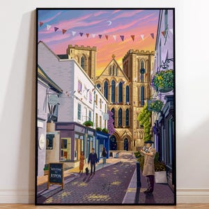 Pode incluir: Uma impressão emoldurada de uma cena de rua em Ripon, Inglaterra, com uma catedral e lojas. A obra de arte tem uma paleta de cores quentes com um céu de pôr do sol, bandeirolas e figuras na rua. Uma placa diz "Welcome to Ripon".