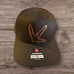 May include: Black trucker hat with a brown bird footprint design on the front. The hat has a mesh back and a black adjustable strap with the text "EST. 1970 RICHARDSON. ORIGINAL 112 TRUCKER ADJUSTABLE" on a white label.