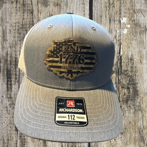 May include: A gray and white trucker hat with a camouflage and American flag patch that says "2nd 1776 Amendment". The hat is adjustable and has a Richardson logo on the back.