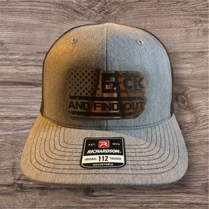 May include: A gray and black trucker hat with a gold patch that says "F*CK AROUND AND FIND OUT" with an American flag design. The hat is adjustable and has a Richardson 112 label.