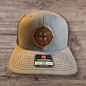 May include: A gray and black trucker hat with a brown leather patch that says "If you can read this you're in range". The hat also has a red and white label that says "Richardson 112 Original Trucker Adjustable".