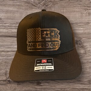 May include: Black trucker hat with a silver metal patch that says "F*CK AROUND AND FIND OUT" with an American flag design. The hat has a black mesh back and a Richardson 112 adjustable strap.