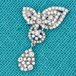 May include: Silver-toned brooch with a butterfly-like design, adorned with numerous clear, sparkling crystals. The brooch features a teardrop-shaped pendant also covered in crystals. Set against a textured teal background.