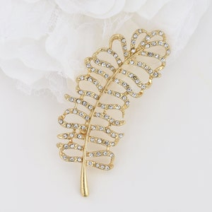 May include: A gold-toned leaf-shaped brooch, adorned with numerous clear, sparkling crystals. The brooch has a detailed, openwork design, with the crystals set along the veins and edges of the leaf. The stem is a solid gold color.
