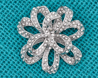 May include: A silver-toned metal brooch shaped like a multi-looped bow or flower, entirely encrusted with small, clear, sparkling rhinestones. The design features several overlapping, curved ribbon-like segments arranged in a symmetrical pattern.