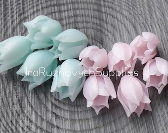 May include: A collection of pastel-colored flower-shaped polymer clay charms. The charms are in shades of light blue and pink.  The charms are arranged in a semi-circle on a wooden surface.