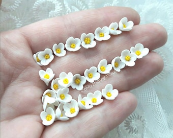 May include: A collection of small, white polymer clay flowers with yellow centres. The flowers are scattered on a hand.