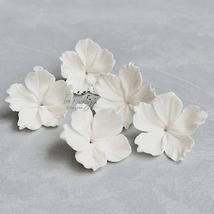 May include: Five white porcelain flower blossoms, each with five petals, arranged on a light grey surface. The flowers are delicate and realistic, with a subtle texture. Ira Ruzhovich Supplies is printed in black on the surface.