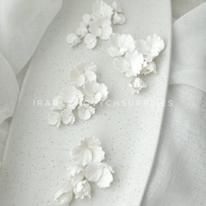 May include: White floral embellishments arranged on a speckled, oval-shaped surface. The delicate flowers are clustered in small groups, with detailed petals. The background includes a white fabric. The text "IRARUZ ZYCHSUPPLIES" is visible.