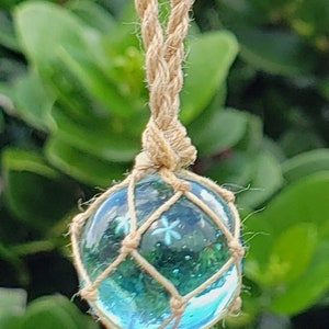 May include: A glass orb earring with a blue and green interior, encased in a woven net and suspended by a natural fiber cord. The earring hangs against a backdrop of green foliage and a white flower.