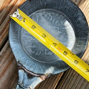 May include: A gray and blue ceramic spoon rest with a handle, measuring approximately 6 inches in diameter. The spoon rest has a speckled pattern and a dark brown rim. A yellow tape measure is placed across the spoon rest.