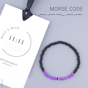 Puede incluir: Una pulsera de cuentas negras con una sección de cuentas moradas. Una etiqueta blanca con el texto "MAKE A WISH" y "11:11" es visible, junto con las palabras "MORSE CODE BRACELET" y una dirección web. También se muestran las palabras "MORSE CODE".