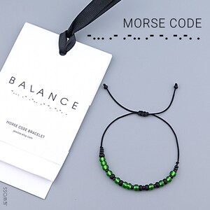 Op de afbeelding: Een zwarte verstelbare armband met groene en zwarte kralen. De armband ligt naast een wit label met het woord "BALANCE" en de tekst "MORSE CODE BRACELET". De afbeelding bevat ook de Morsecode voor "BALANCE".
