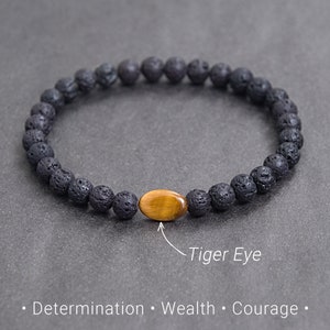 Könnte beinhalten: Schwarzes Lava-Gestein-Perlenarmband mit einem ovalen Tigerauge-Stein in der Mitte. Das Armband ist elastisch. Das Bild enthält den Text "Tiger Eye" und die Wörter "Entschlossenheit, Reichtum, Mut."
