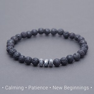 Könnte beinhalten: Ein schwarzes Lavastein-Armband mit drei silberfarbenen Perlen. Das Armband besteht aus kleinen, runden, porösen Perlen. Der Hintergrund ist einfarbig grau. Der Text am unteren Bildrand lautet "Calming • Patience • New Beginnings."