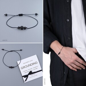 Peut inclure: Bracelet en pierre de lave noire avec cordon réglable. Le bracelet comporte trois grosses perles de lave et deux petites perles. La carte à côté du bracelet indique "Grounding" et énumère des bienfaits tels que le calme et la force.