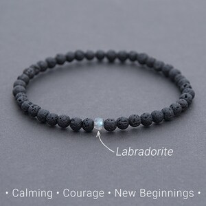 Könnte beinhalten: Schwarzes Lava-Stein-Armband mit einer einzelnen schillernden Labradorit-Perle. Das Armband liegt auf einer grauen Oberfläche und der Text "Calming. Courage. New Beginnings." befindet sich unterhalb des Armbands.